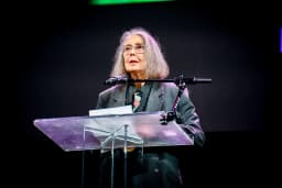 Anne Carson, in glasses and a grey jacket, speaks at a clear acrylic lectern at the 2024 National Book Awards finalist reading in New York.