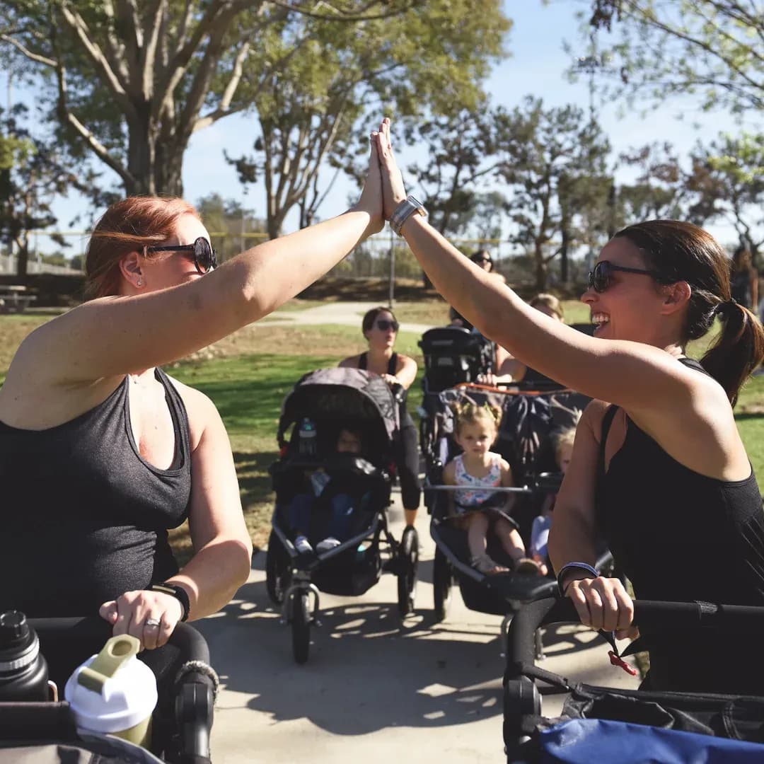 FIT4MOM moms giving high fives while pushing strollers at a Stroller Strides class in a sunny park