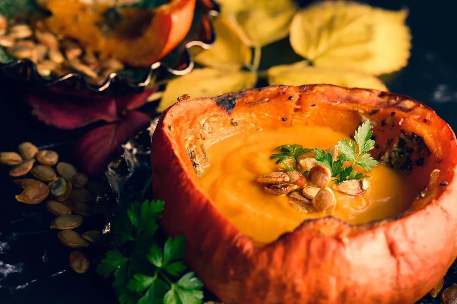 Pumpkin soup served inside a hollowed-out pumpkin, topped with toasted pumpkin seeds and herbs, surrounded by autumn leaves.