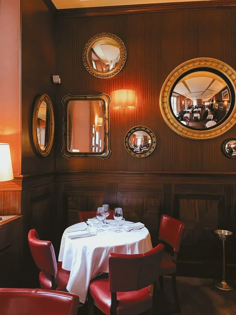 A wood-panelled dining room with round gilt mirrors, red leather chairs and a table laid with a white cloth.