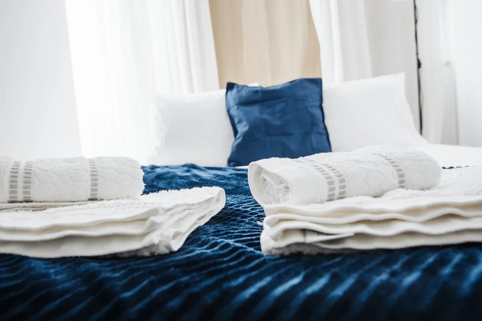 Folded white towels laid out on a freshly made bed with a blue velvet cover and cushion.
