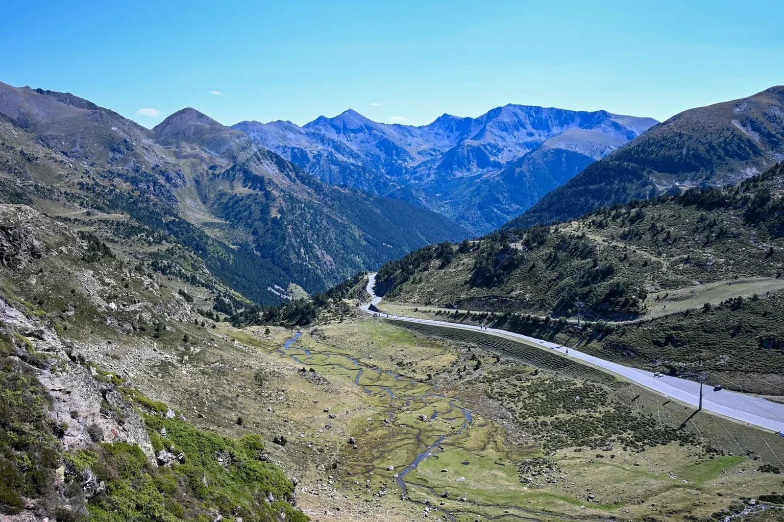A road winding through a green valley beneath high mountain peaks under a blue sky.