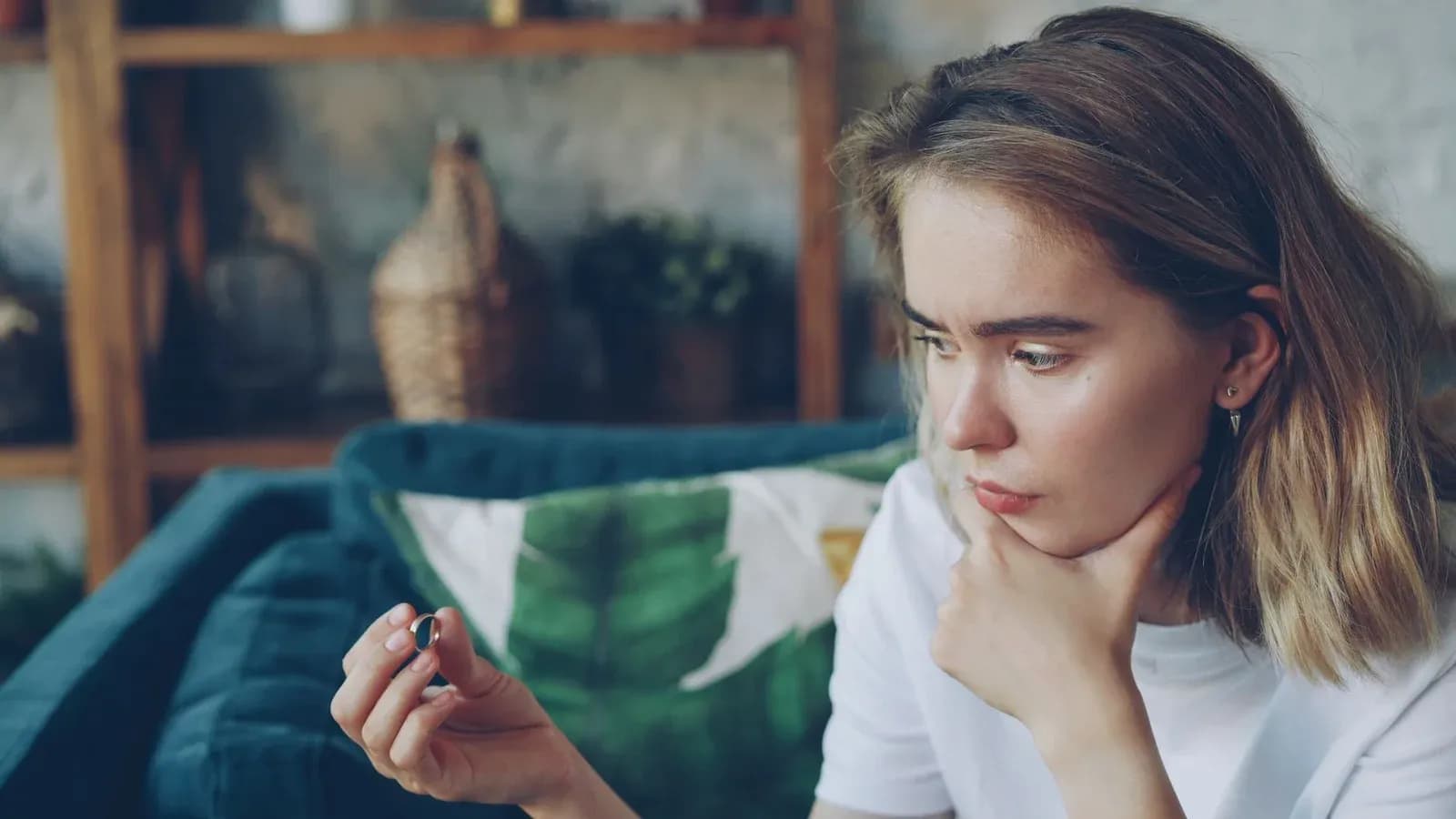 A young woman sits on a sofa, chin in hand, looking closely at a small object as she weighs a decision.