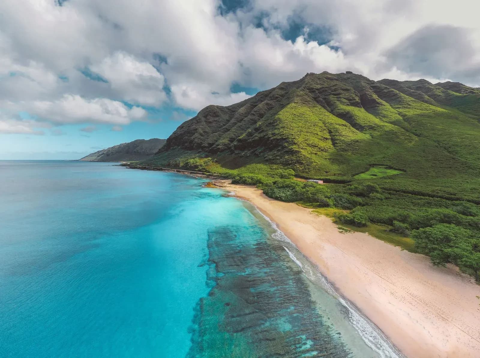 A turquoise bay curves along a white sand beach below green mountains on the Hawaiian coast.