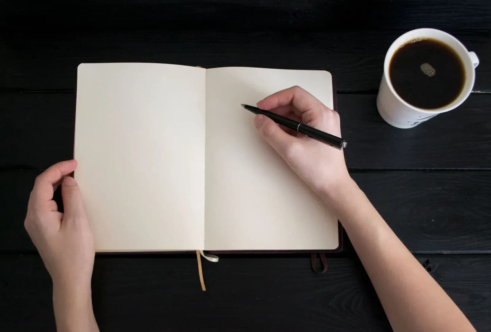 A hand writes in an open notebook beside a cup of black coffee on a dark table.