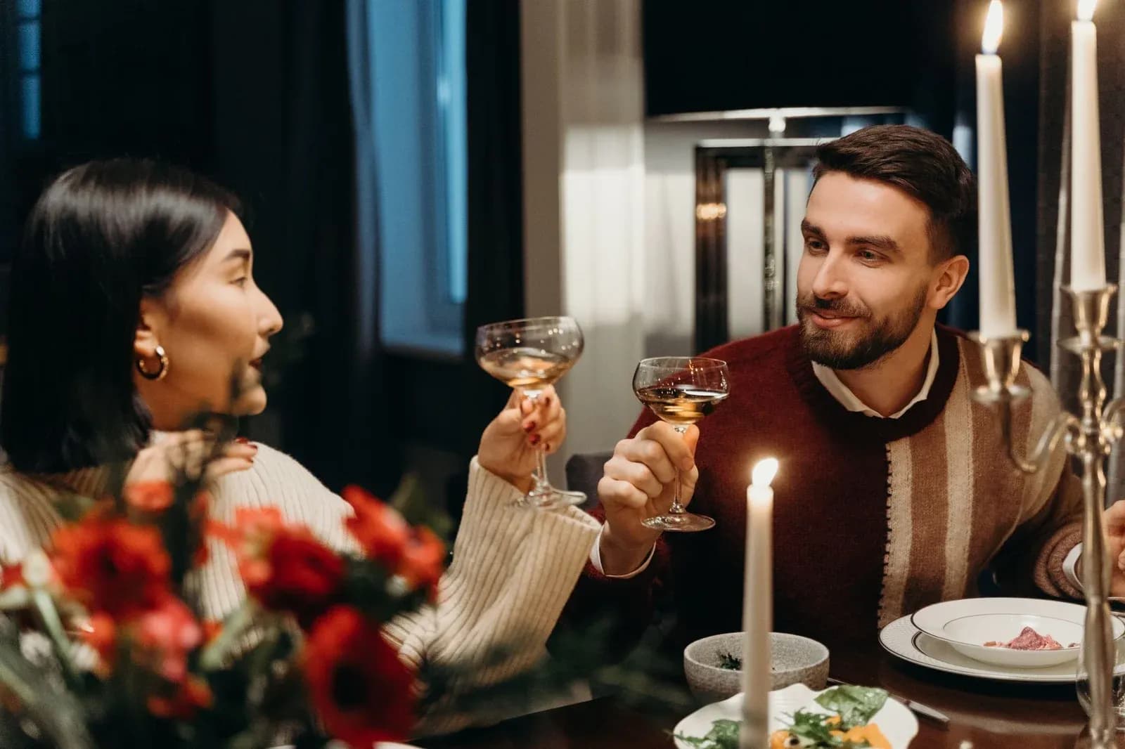 A couple raise their glasses to each other across a candlelit dinner table.