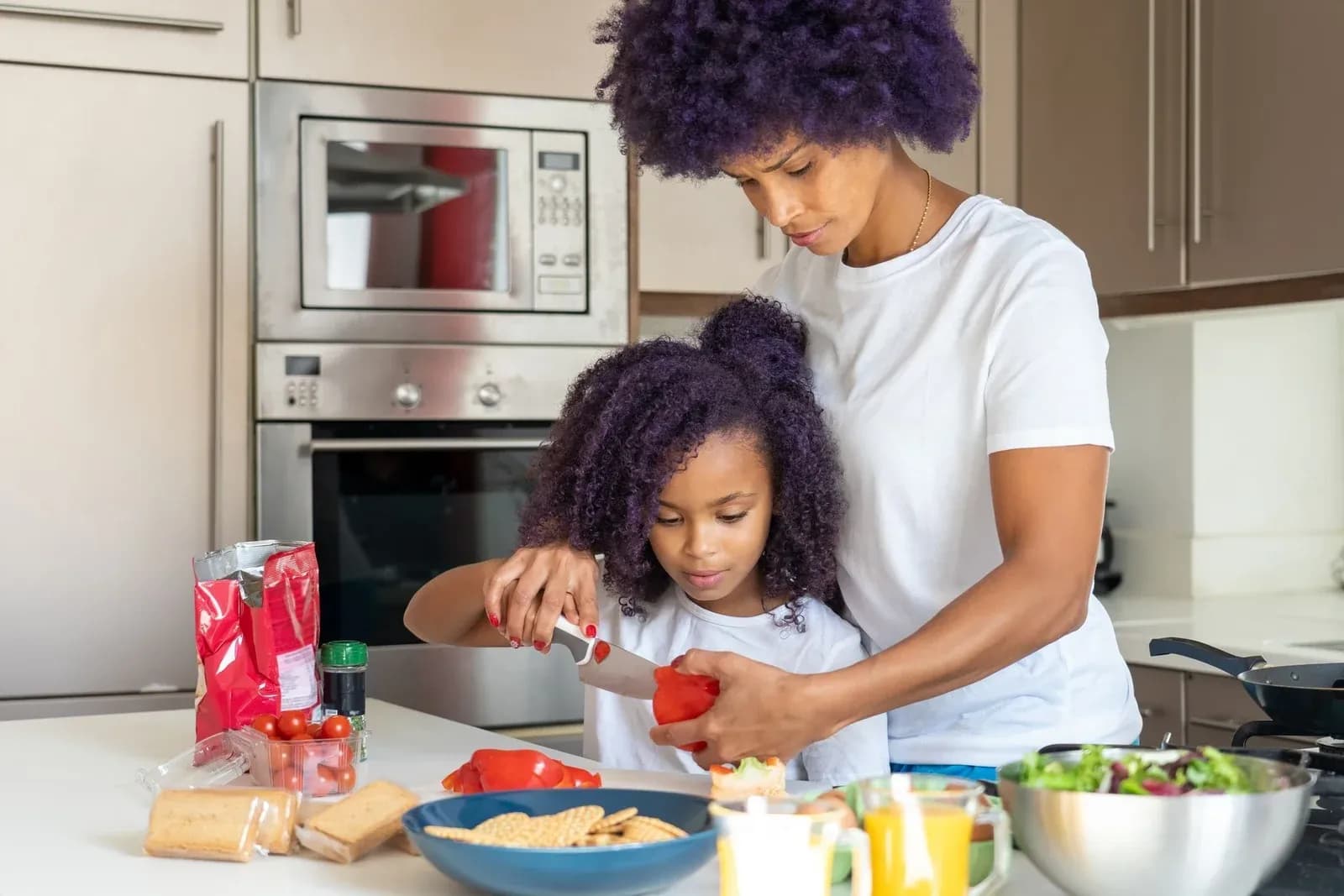 A mother helps her young daughter slice red peppers in a home kitchen.