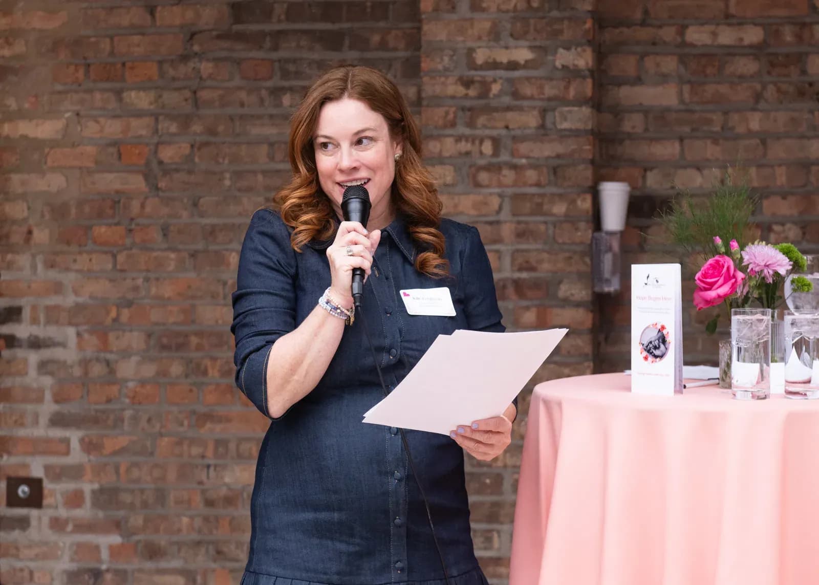 A woman in a denim shirt dress speaks into a handheld microphone while holding printed notes, standing beside a table with a pink cloth, fresh flowers and a charity brochure in front of an exposed brick wall.