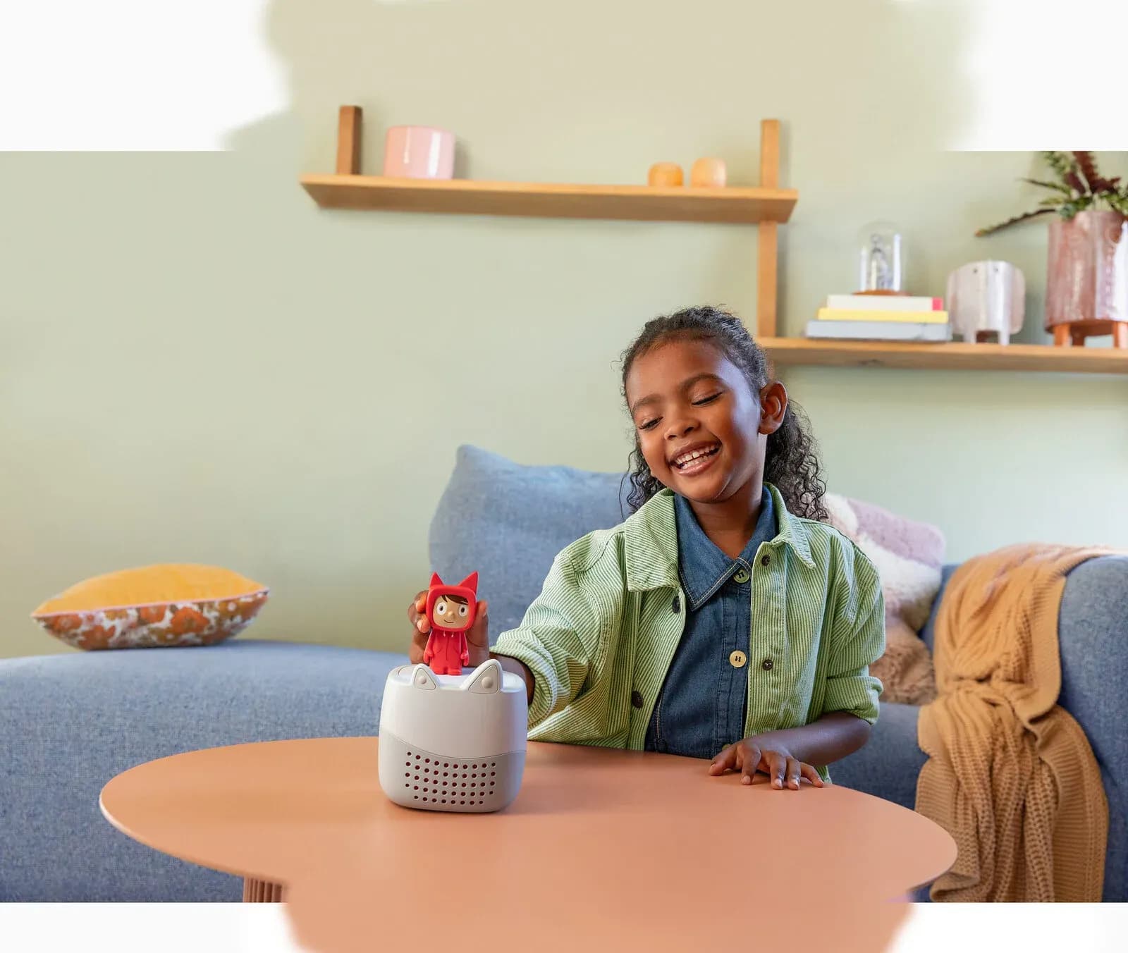 A young child places a red Tonie figure on top of a grey Toniebox Lite on a living room table