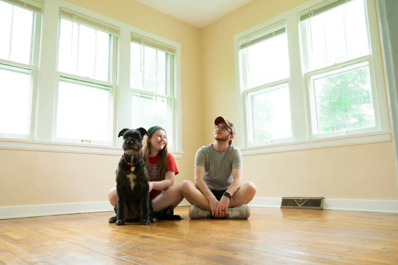 A young couple sits on the wooden floor of an empty, sunlit room with their black dog beside them.