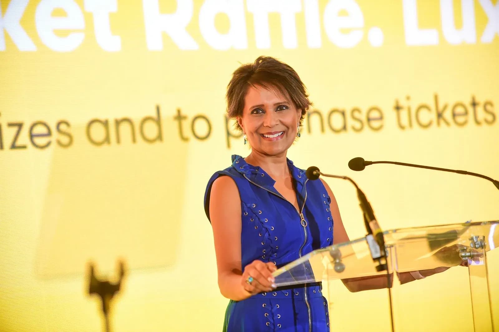 Arti Halai, executive coach and former ITV news presenter, smiling at a clear lectern with two microphones, in a blue sleeveless dress, in front of a yellow screen