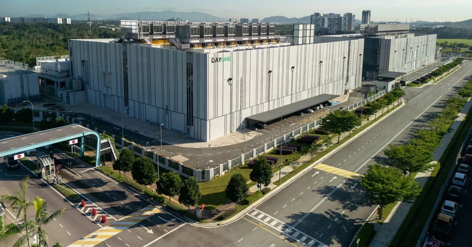 Aerial view of a large grey data centre building with DayOne signage and rooftop cooling units, beside a gated entrance and a tree-lined multi-lane road
