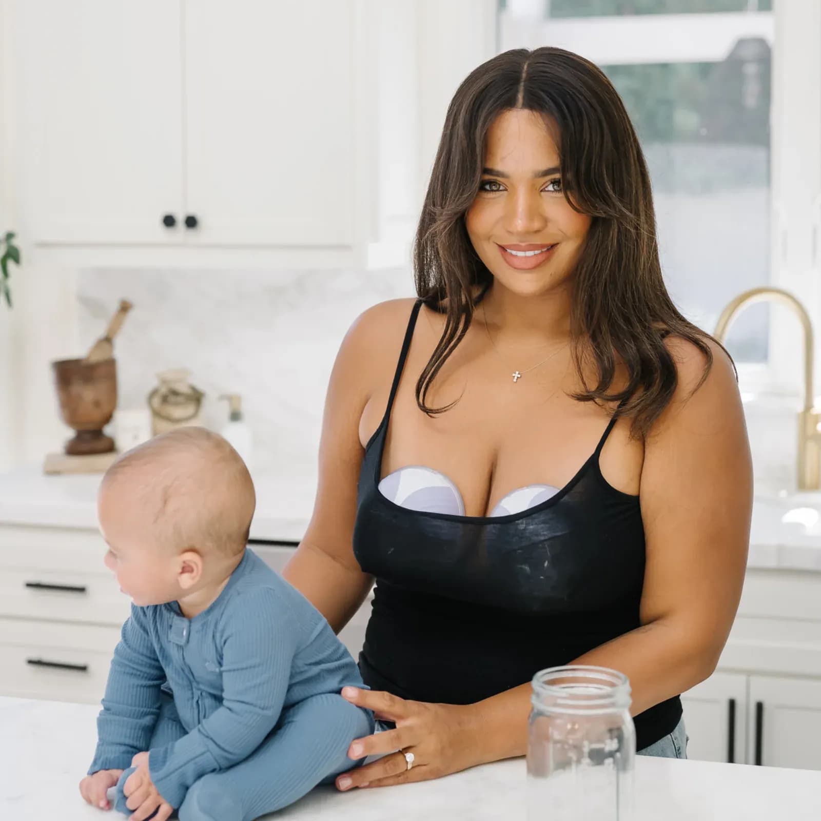 A smiling mother wearing a hands-free BabyBuddha breast pump under her top in a bright kitchen, with her baby sitting on the counter beside her.