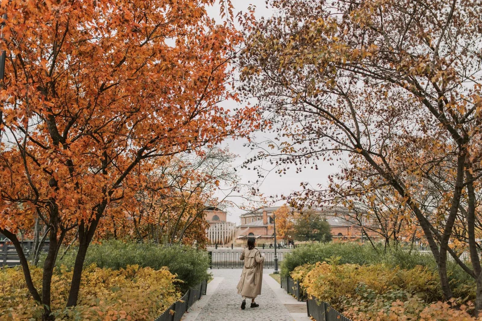 A woman in a beige trench coat walks away from the camera along a path lined with orange and red autumn trees, a red-brick building visible in the distance