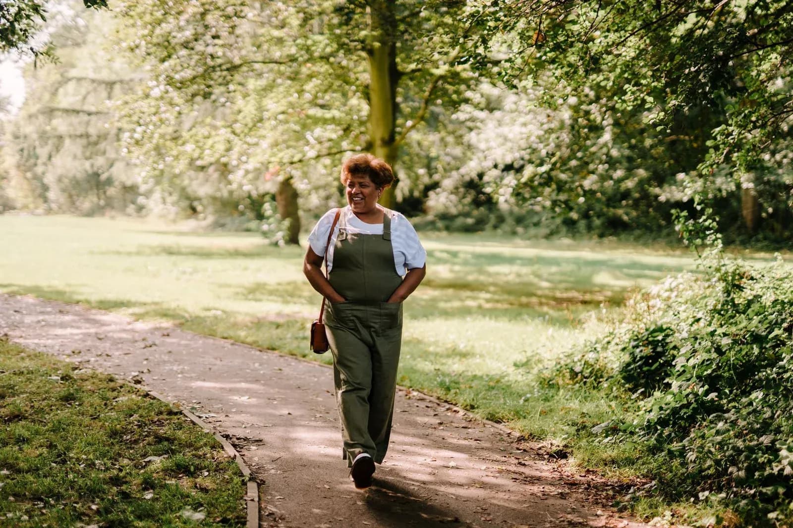 A smiling woman in dungarees walks along a sunlit path through a green park.