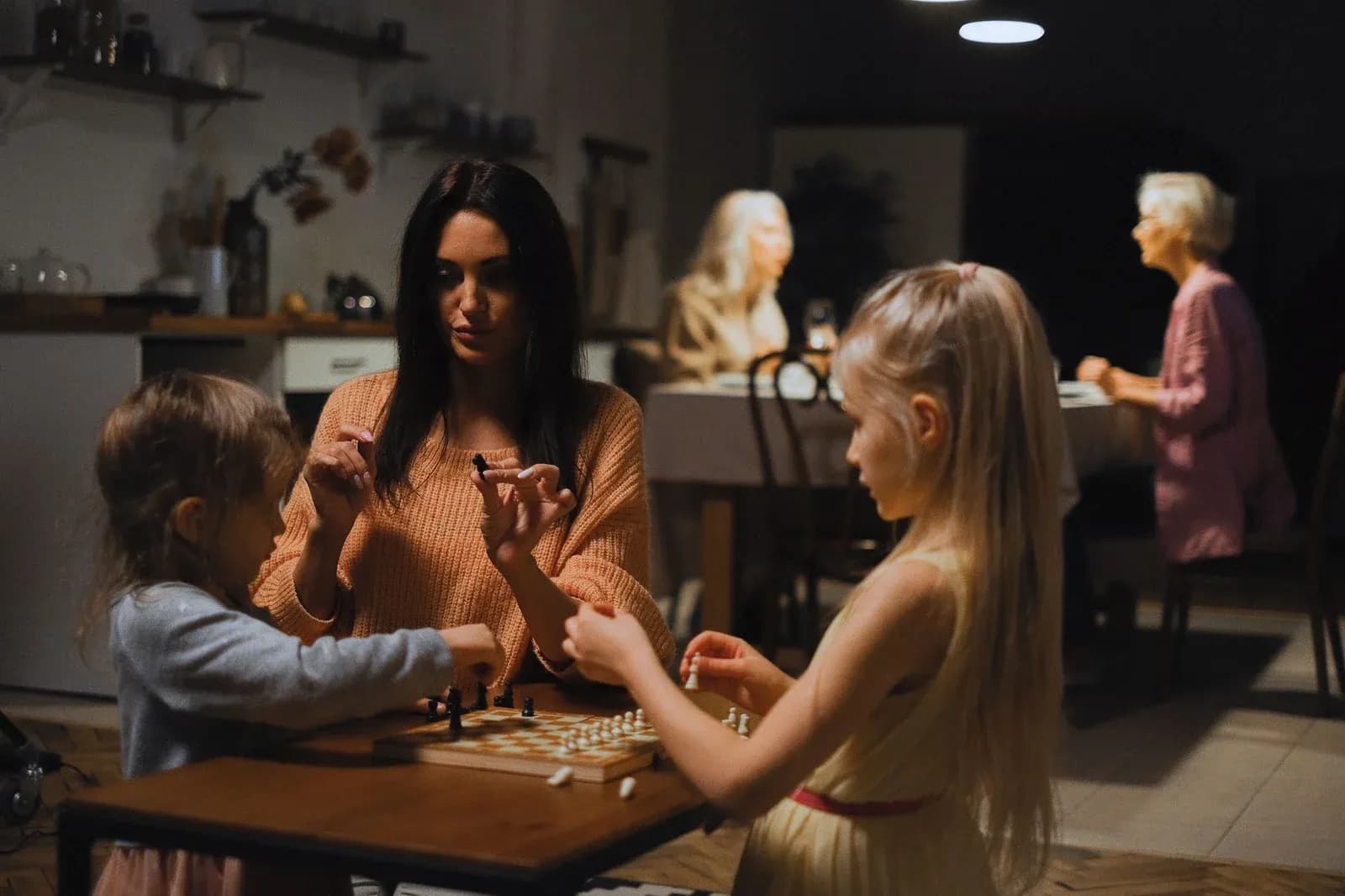 A mother and two daughters play a board game at a kitchen table in the evening.