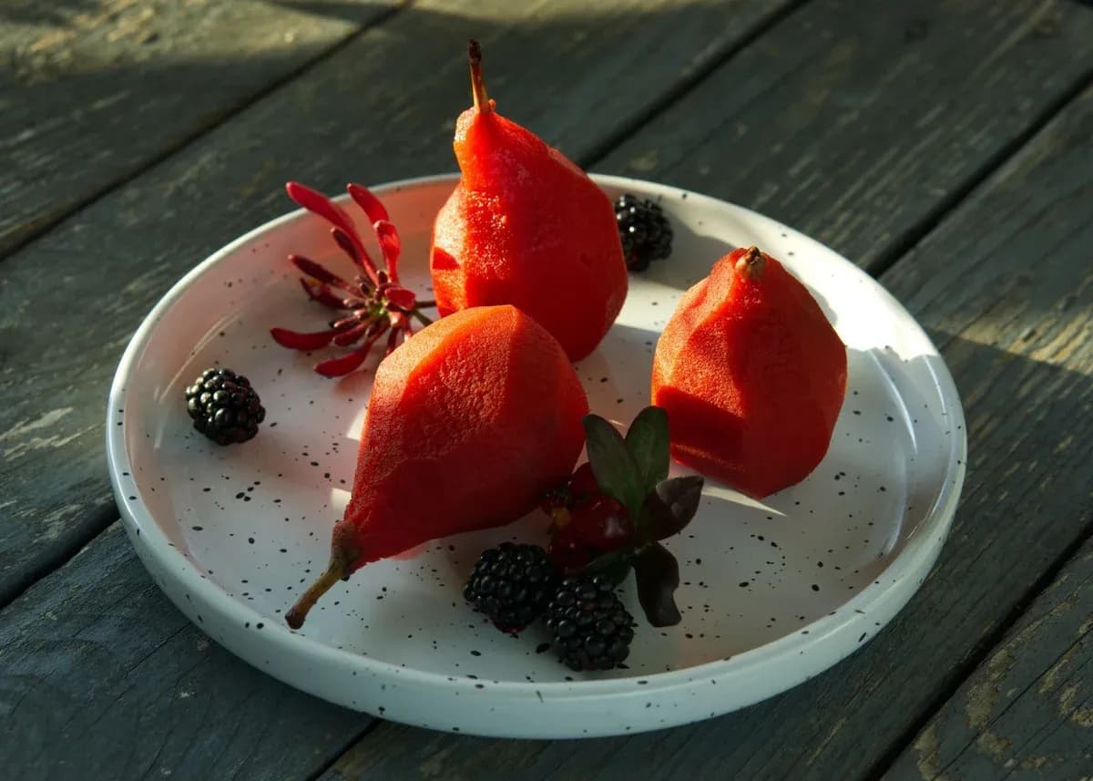 Three ruby-red poached pears on a white plate with blackberries and a red flower.