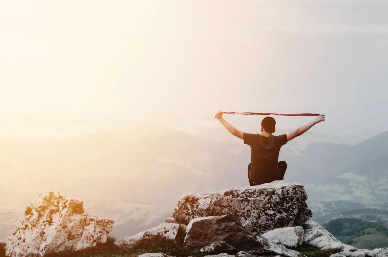 A person sits on a rocky summit above the mountains at sunrise, stretching a band overhead with both arms.