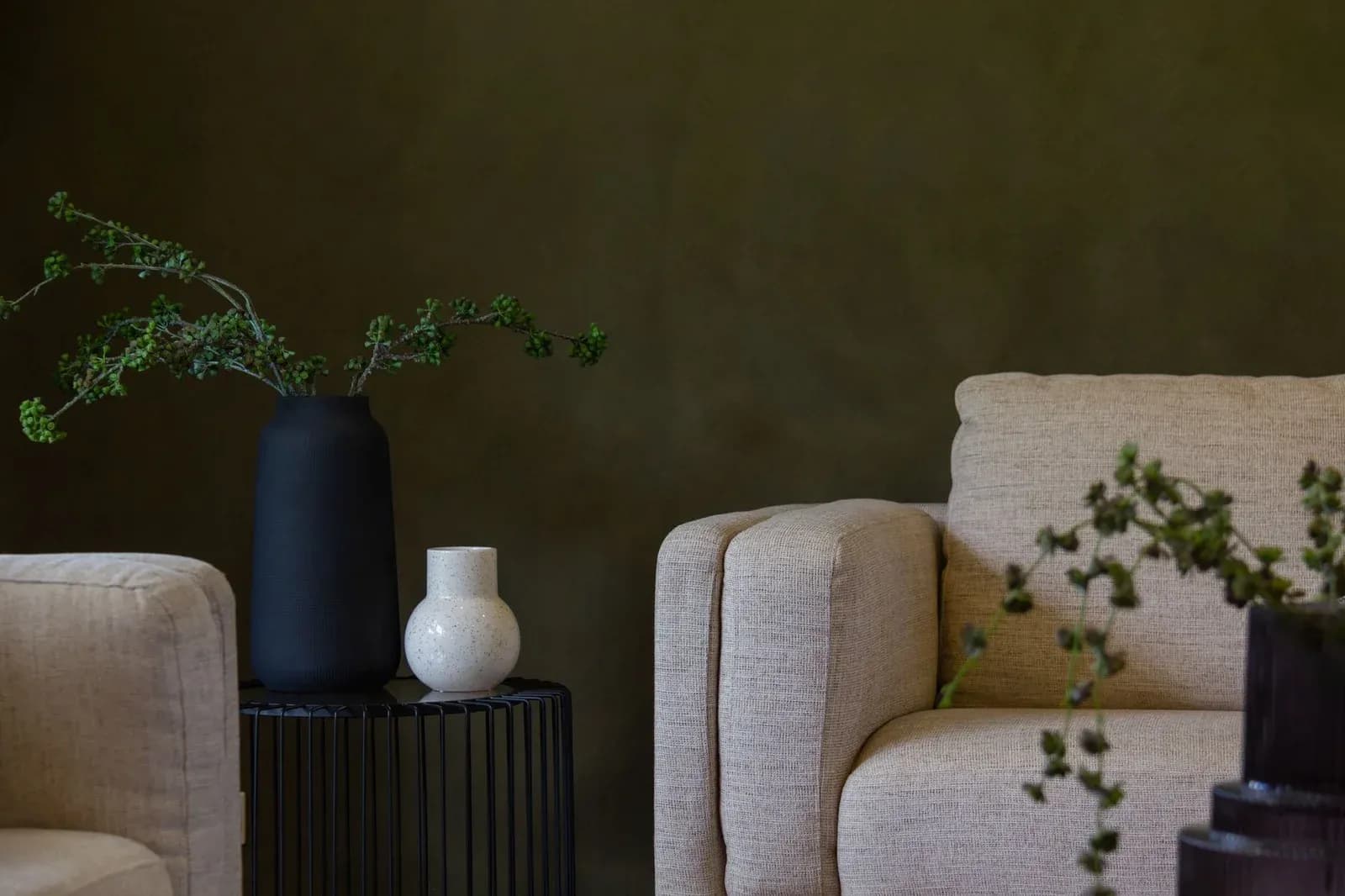 A living room with an olive green painted wall, cream armchairs and a vase of greenery.