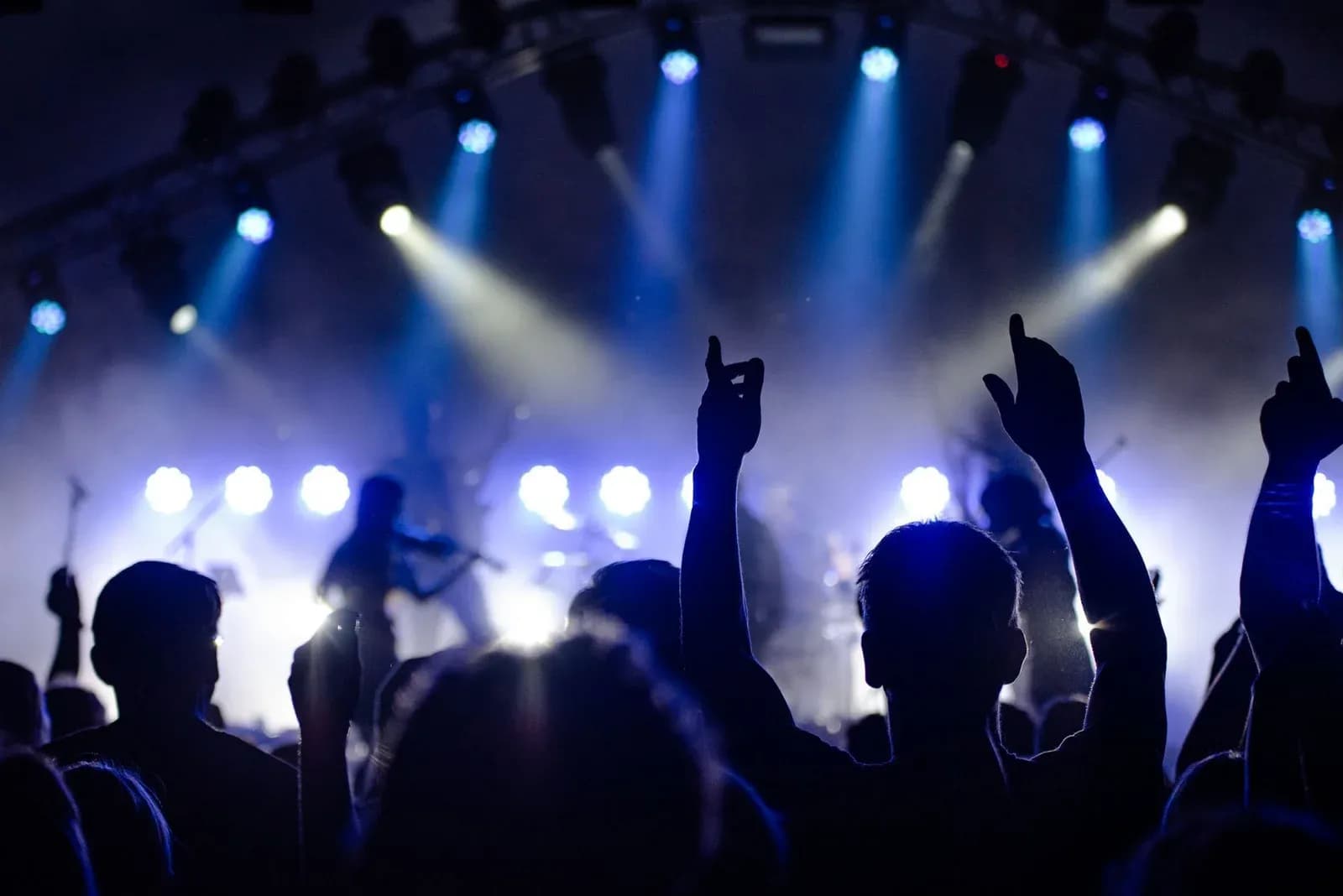 Fans with raised hands in front of a stage lit by beams of blue light at a concert.