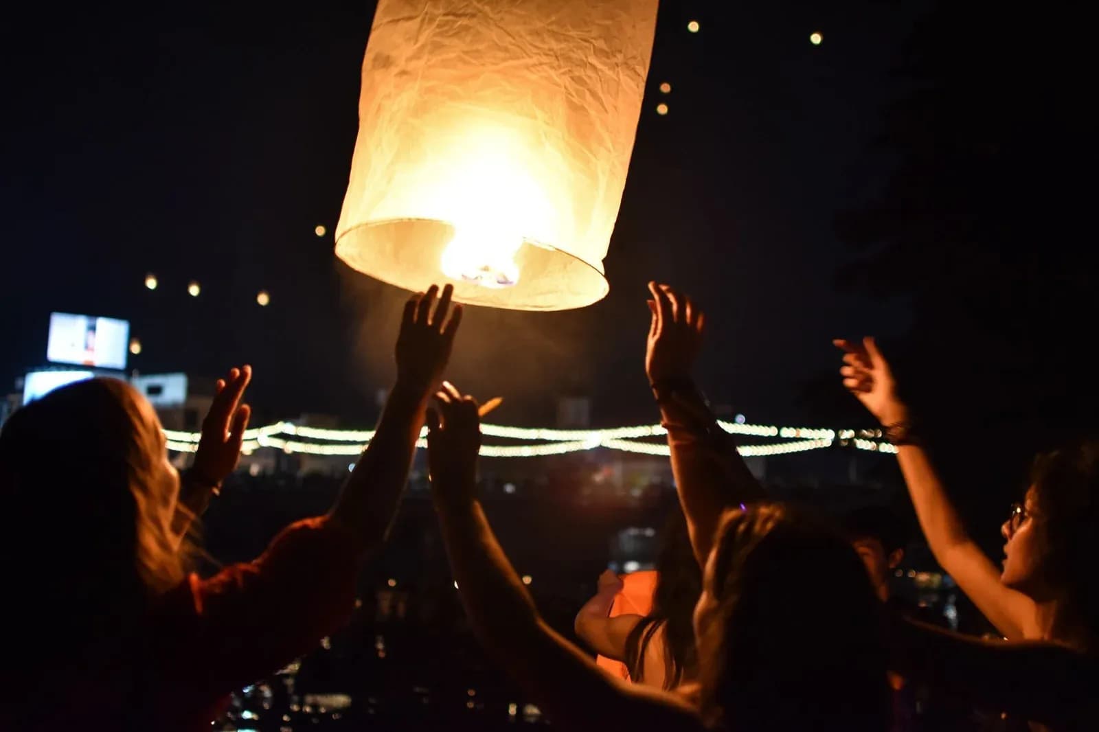 Several women reach up together towards a glowing paper lantern rising into the night sky.