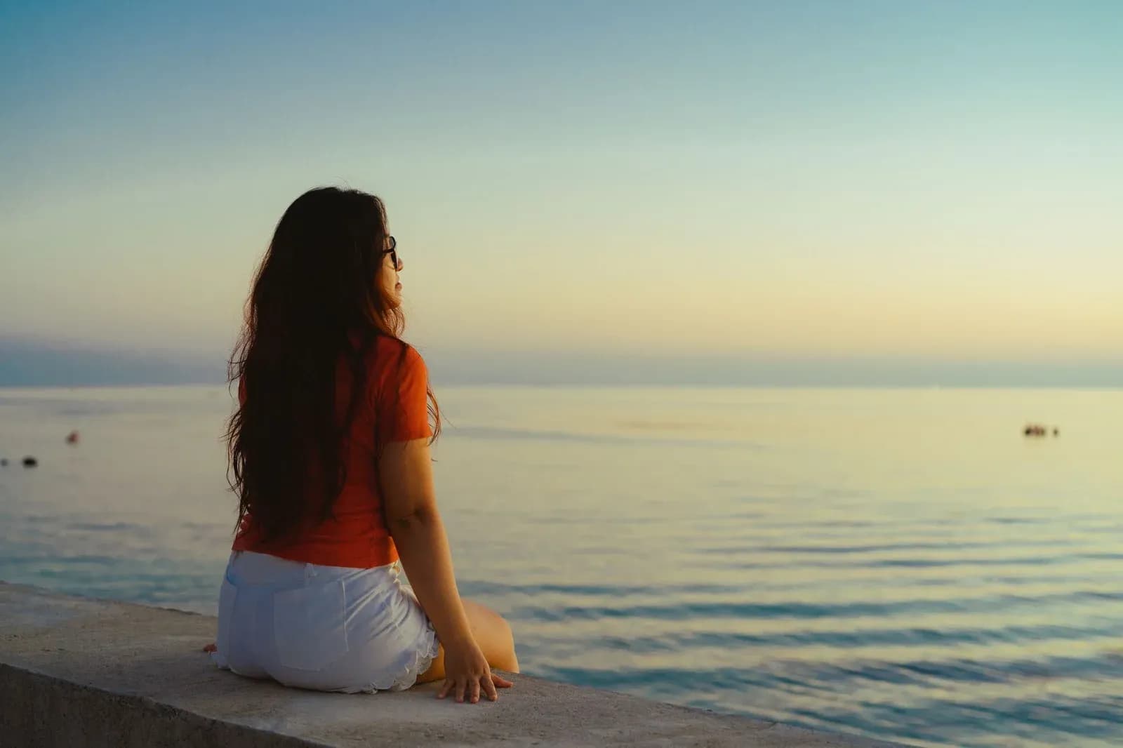 A young woman sits on a sea wall looking out at a calm sea at dawn.