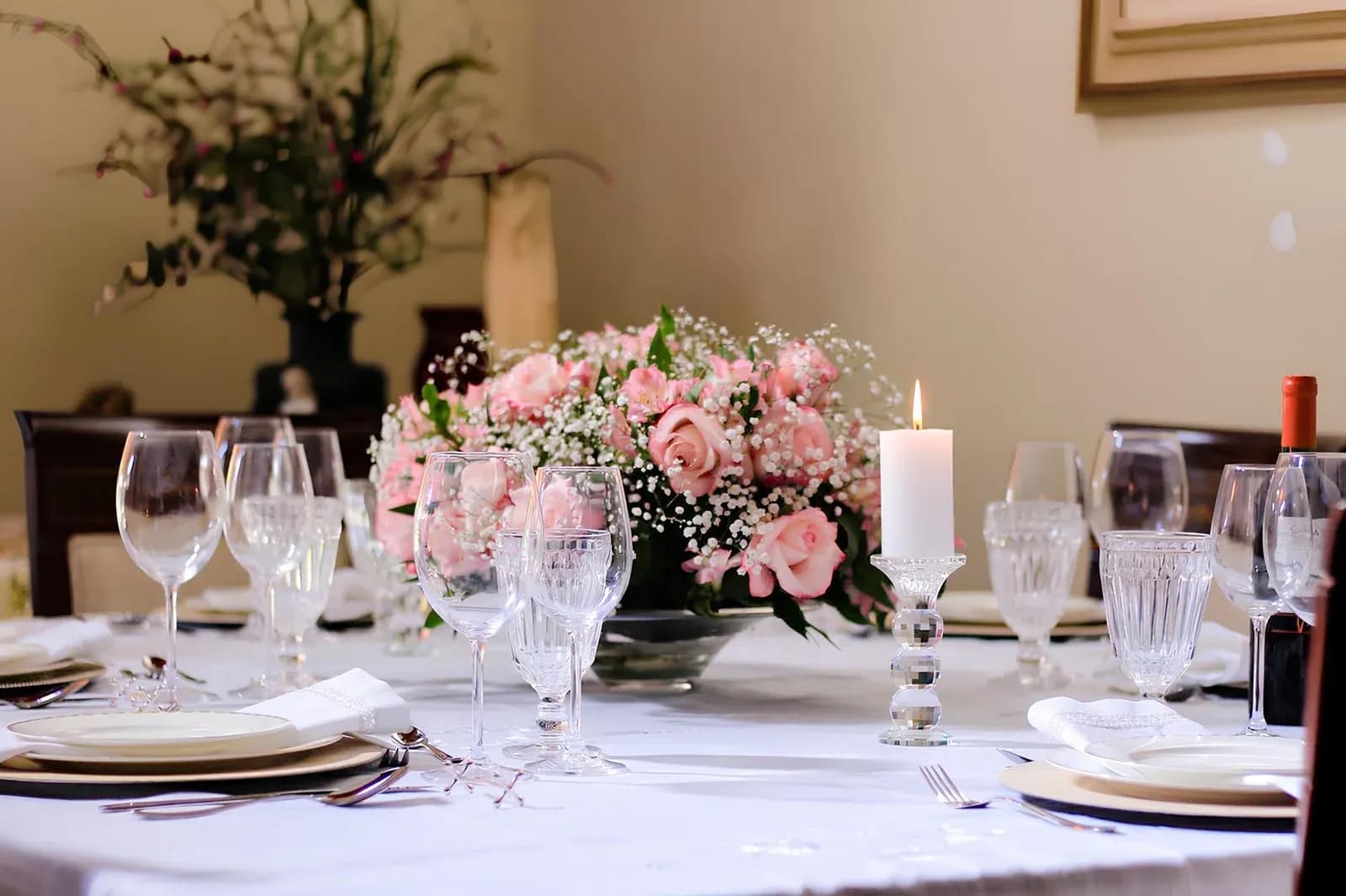 An elegant dinner table laid with white linen, crystal glasses, a lit candle and a centrepiece of pink roses.