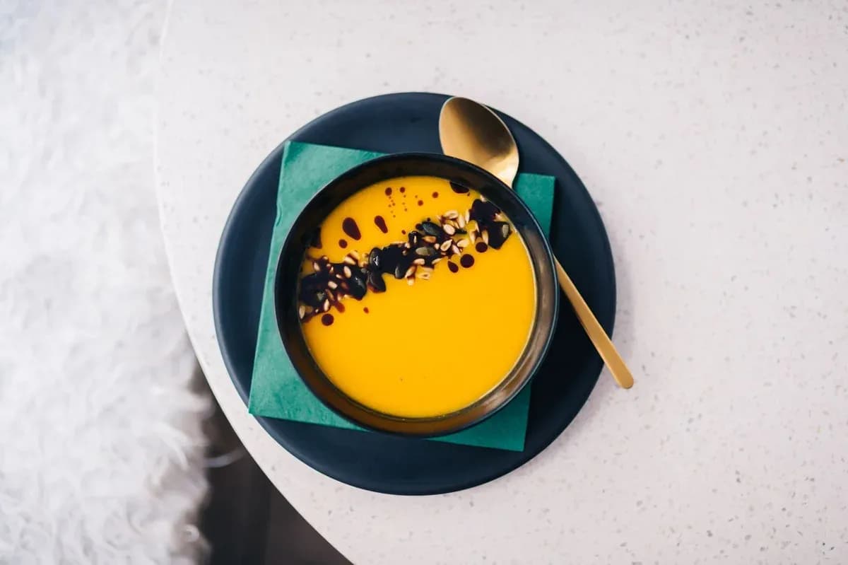A bowl of golden soup topped with seeds, served on a dark plate with a gold spoon.