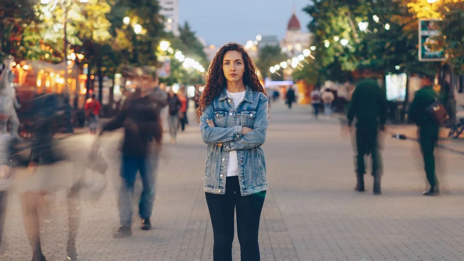 A young woman stands still with her arms crossed in a city street at dusk while the crowd moves past her in a blur.
