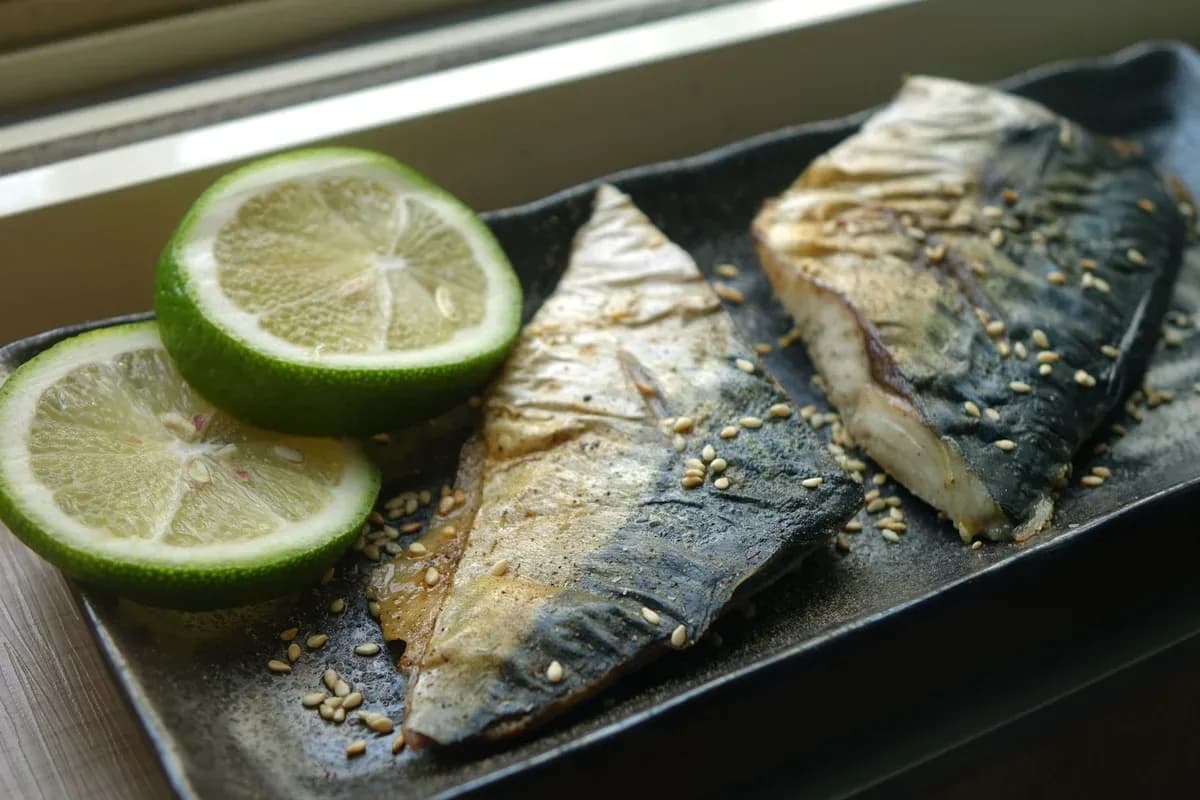 Two grilled mackerel fillets on a dark platter beside halves of lime.