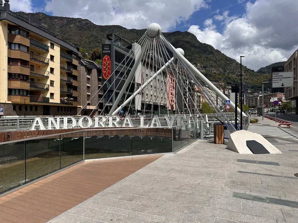 Large white letters spelling Andorra la Vella on a riverside walkway beside a modern bridge, with mountains behind.