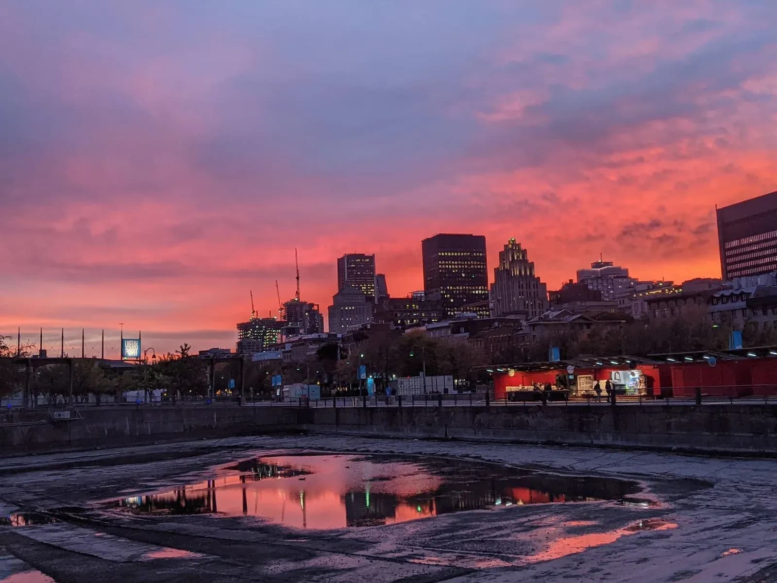 The Montreal skyline and Old Port under a vivid pink and red sunset, reflected in water on the waterfront.