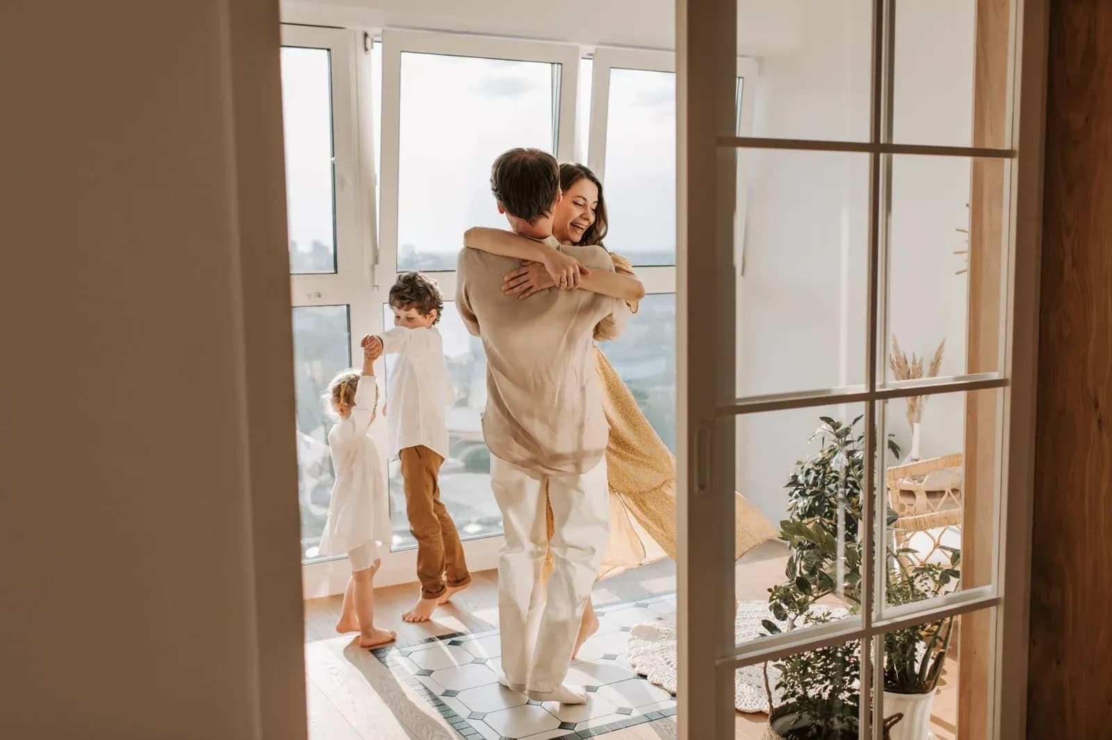 A family dances together in a sunlit living room, seen through an open glass door.