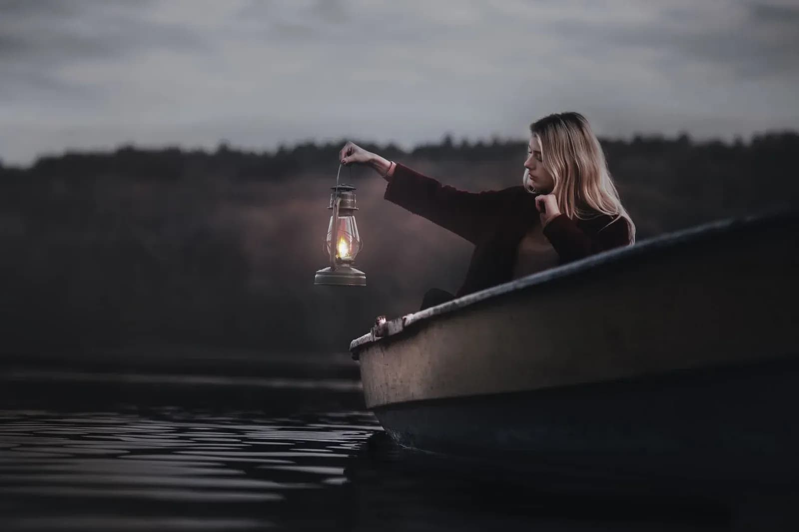 A woman sitting in a boat at dusk holds a lit lantern out over dark water.