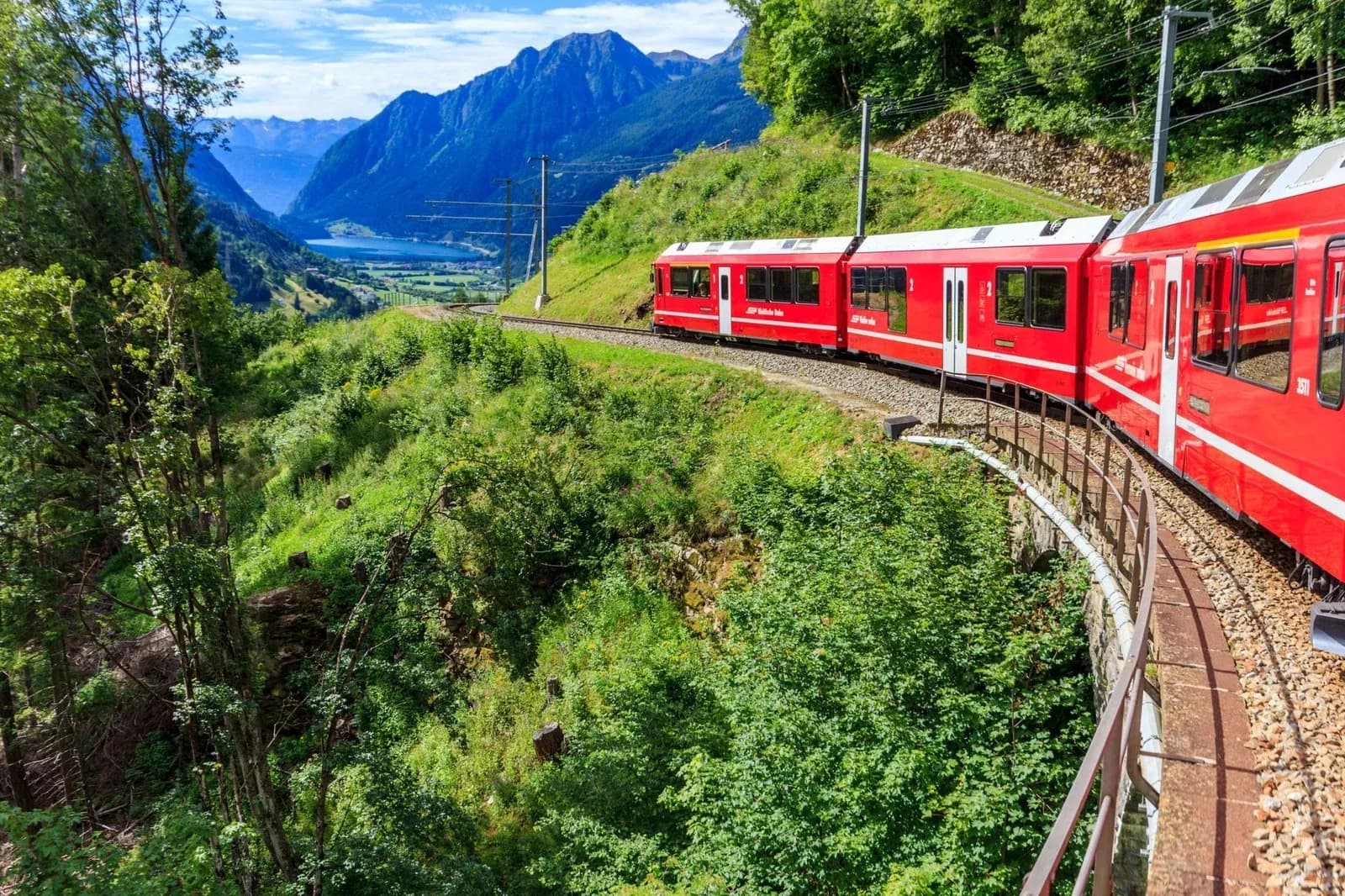 A red mountain train curves along a green hillside above a valley lake, with Alpine peaks behind.