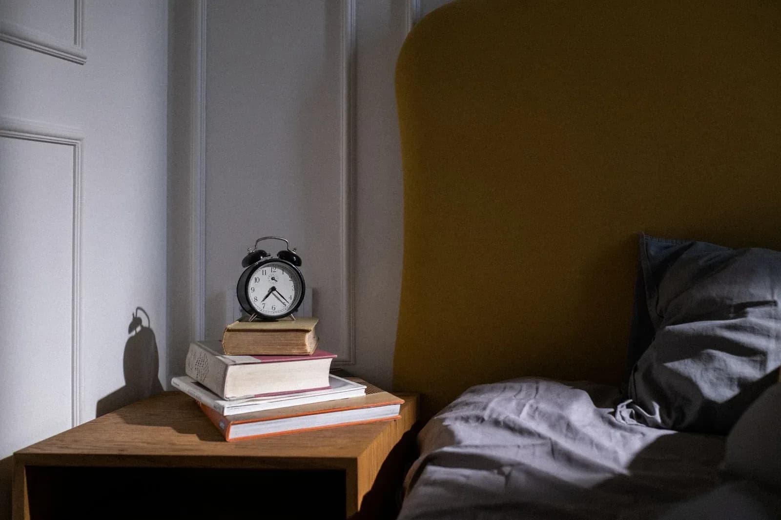 An alarm clock on a stack of books on a bedside table next to an unmade bed in soft early light.