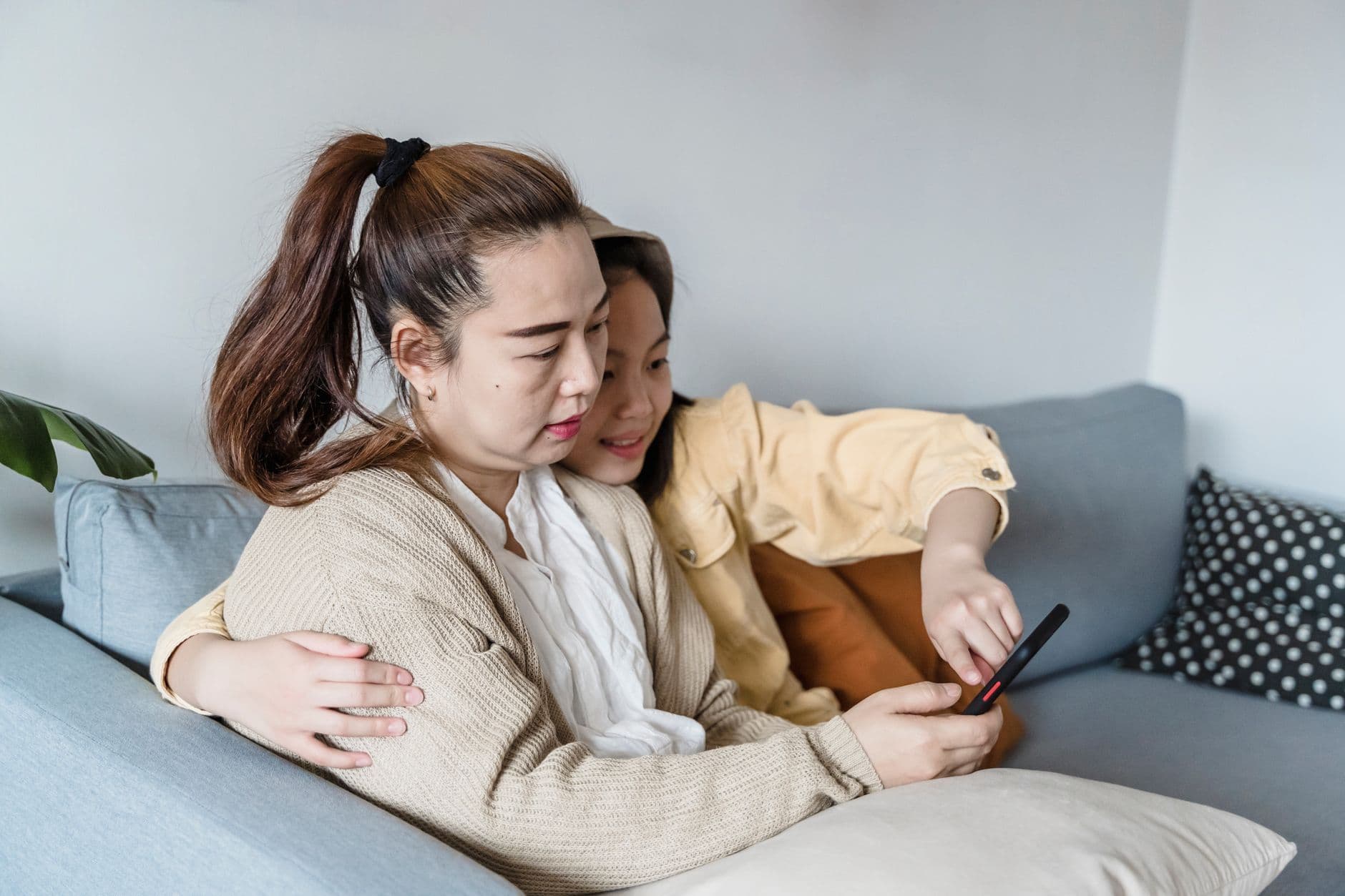 A mother and her daughter sit close together on a sofa at home, looking at a phone
