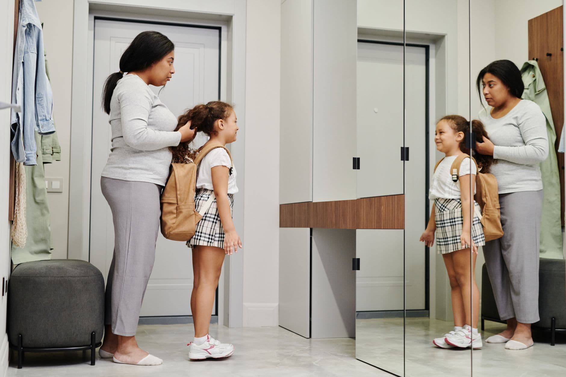 A mother stands behind her school-age daughter, who wears a backpack, as they get ready in front of a hallway mirror