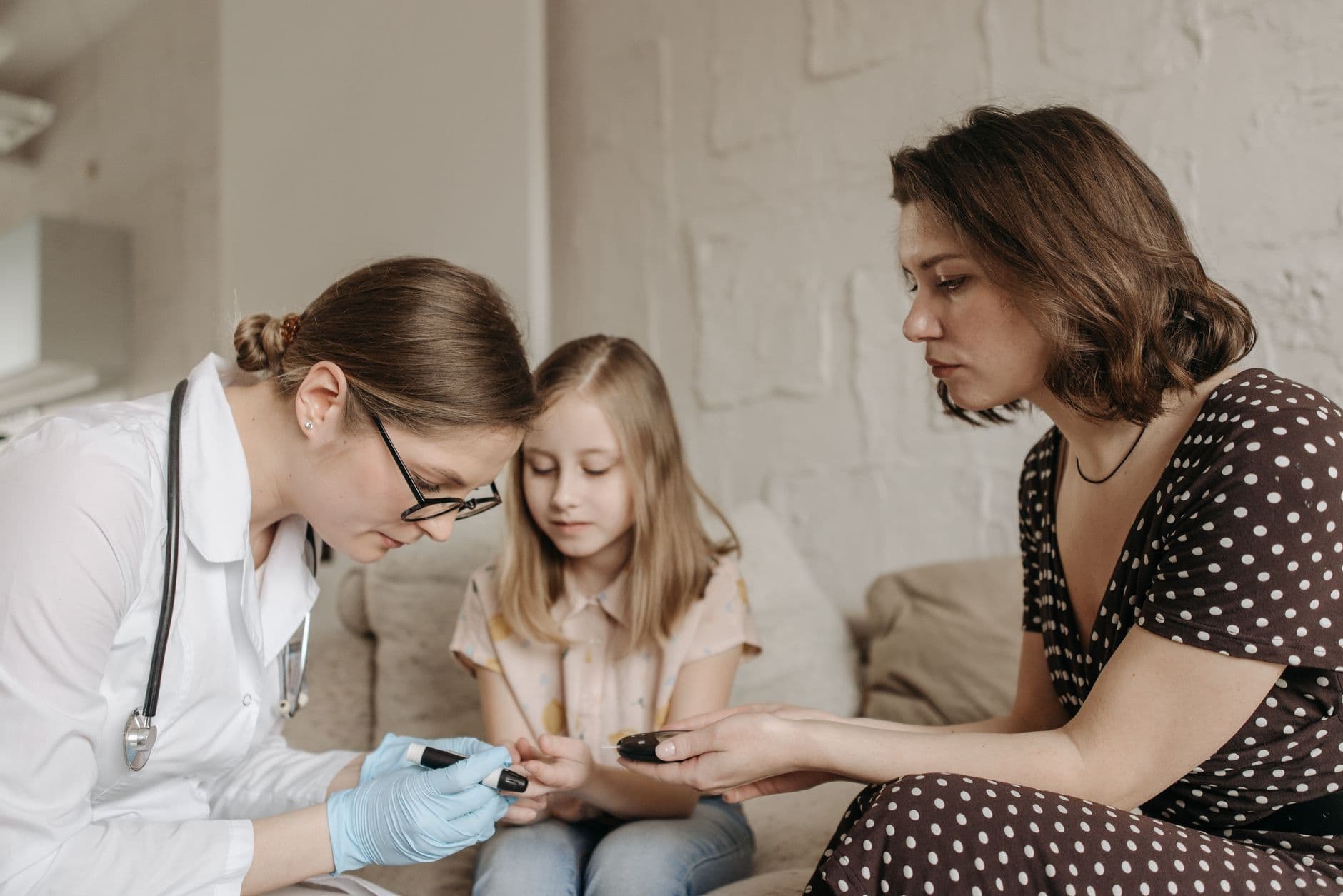 A doctor does a finger-prick blood test on a young girl while her mother holds her hand at home