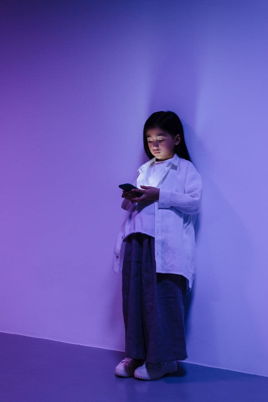 A young girl is engrossed in using a smartphone while standing against a wall with purple lighting.