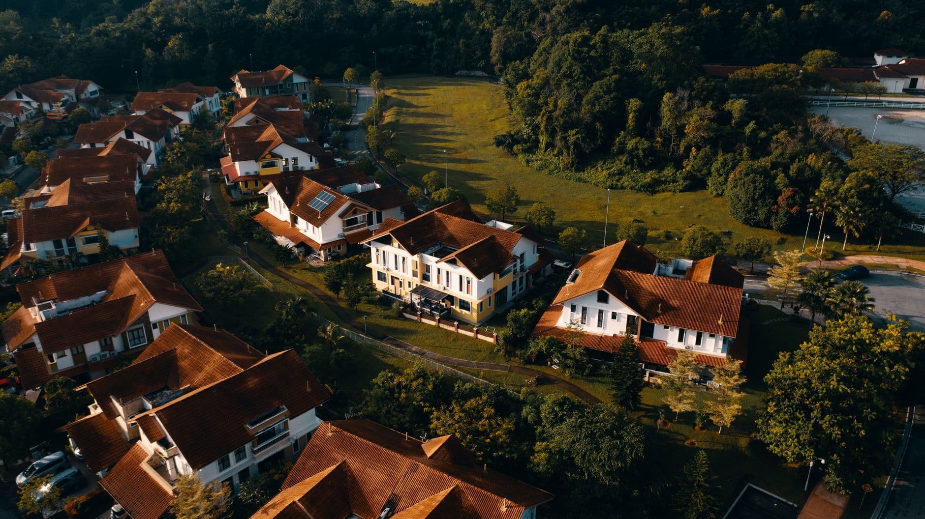 Aerial view of suburban houses with red roofs surrounded by greenery at sunset.