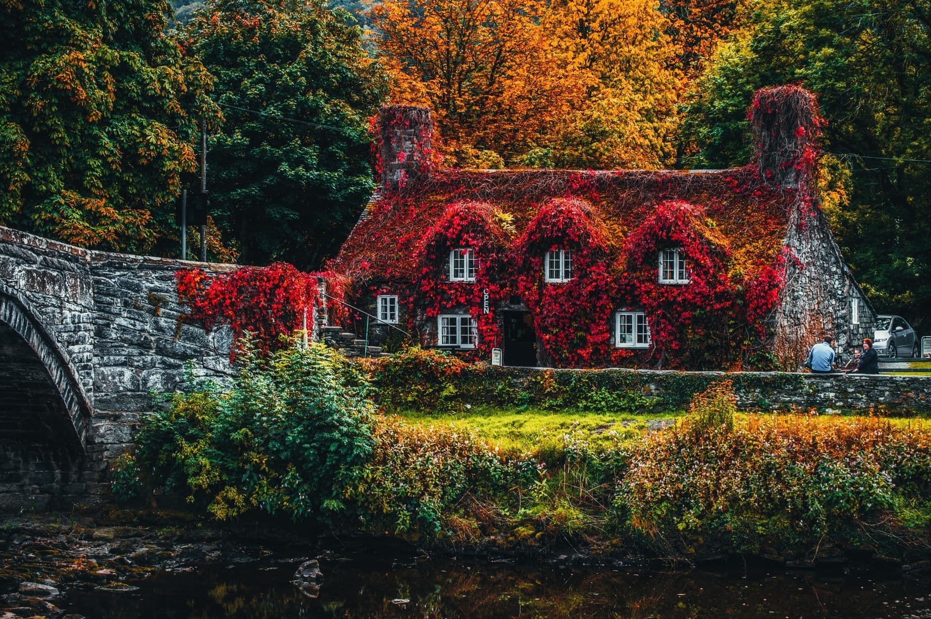 Charming ivy-covered cottage by a stone bridge surrounded by vibrant fall foliage and a peaceful river.