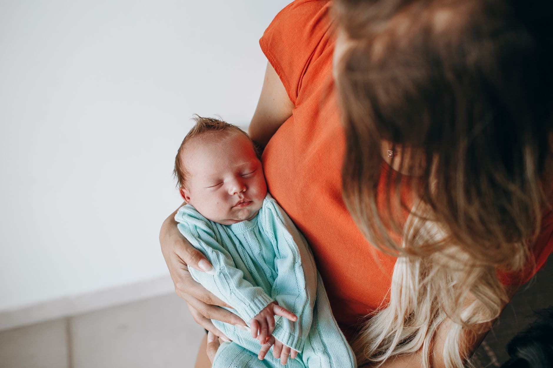 A mother seen from behind holding a sleeping newborn in a mint knit outfit at home