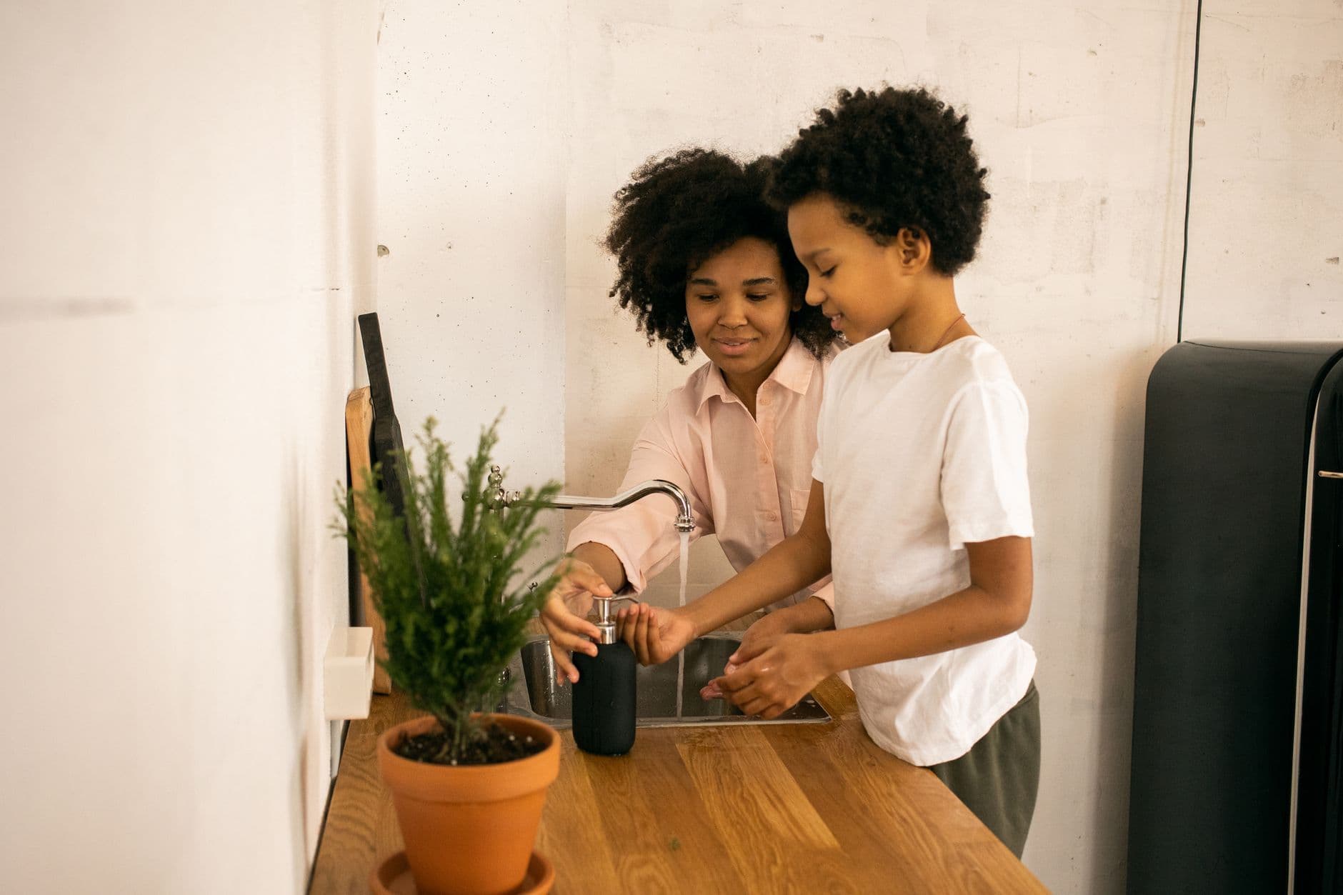 A mother and her young son wash their hands together with soap at a kitchen sink