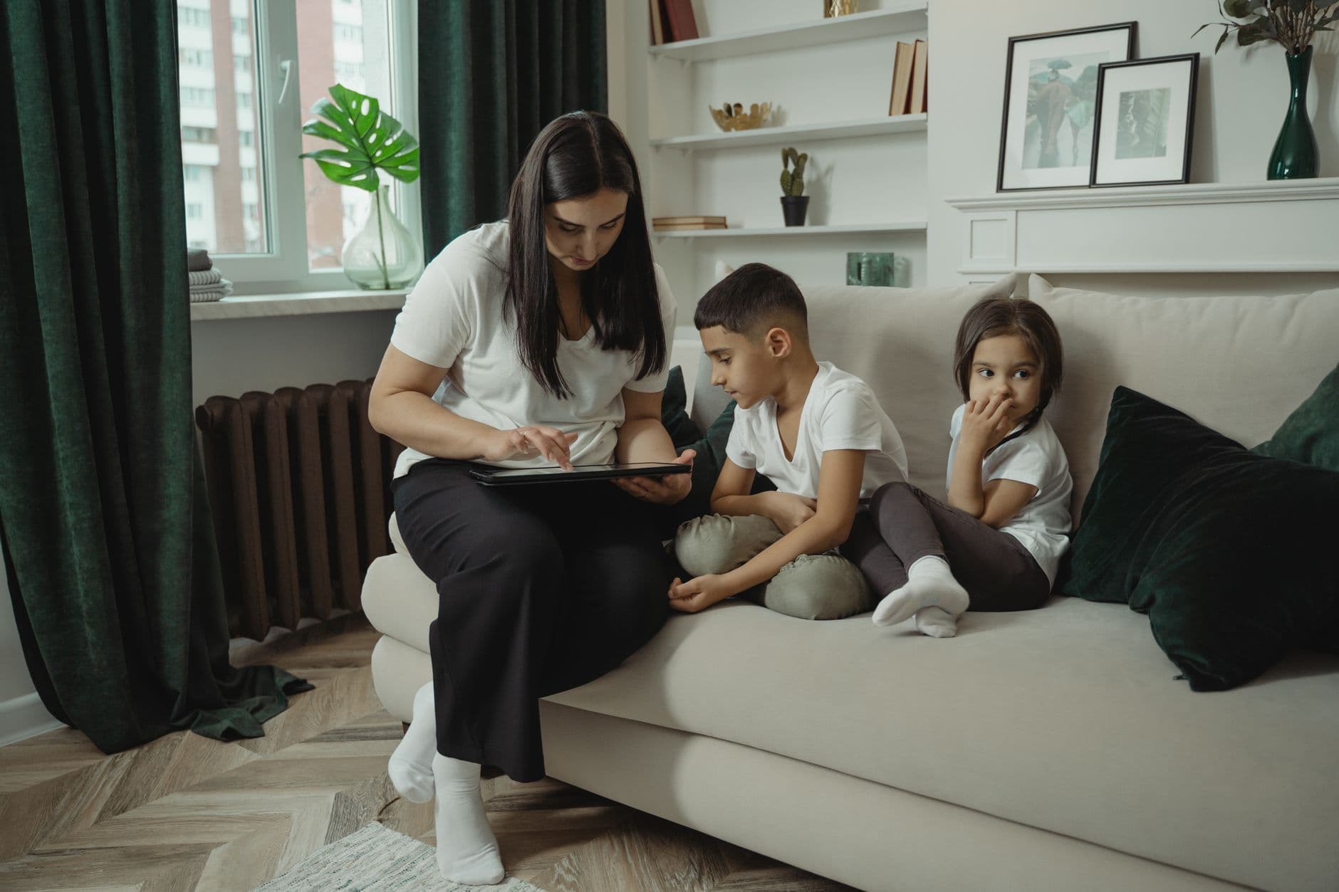 A mother sits on a sofa using a tablet while her son and daughter look at the screen beside her