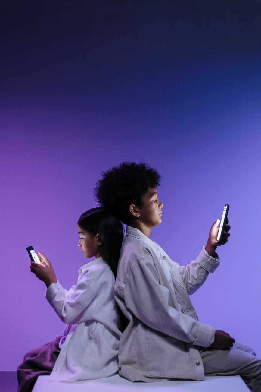 Two children using smartphones while sitting back to back in colorful lighting, highlighting digital engagement.
