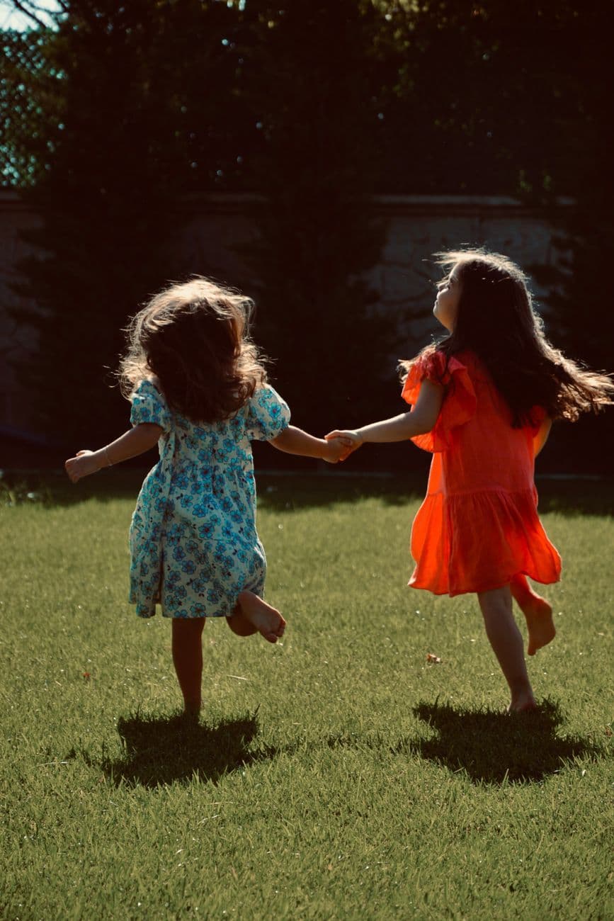 Two girls in vibrant dresses holding hands and running on a sunny lawn, back view.