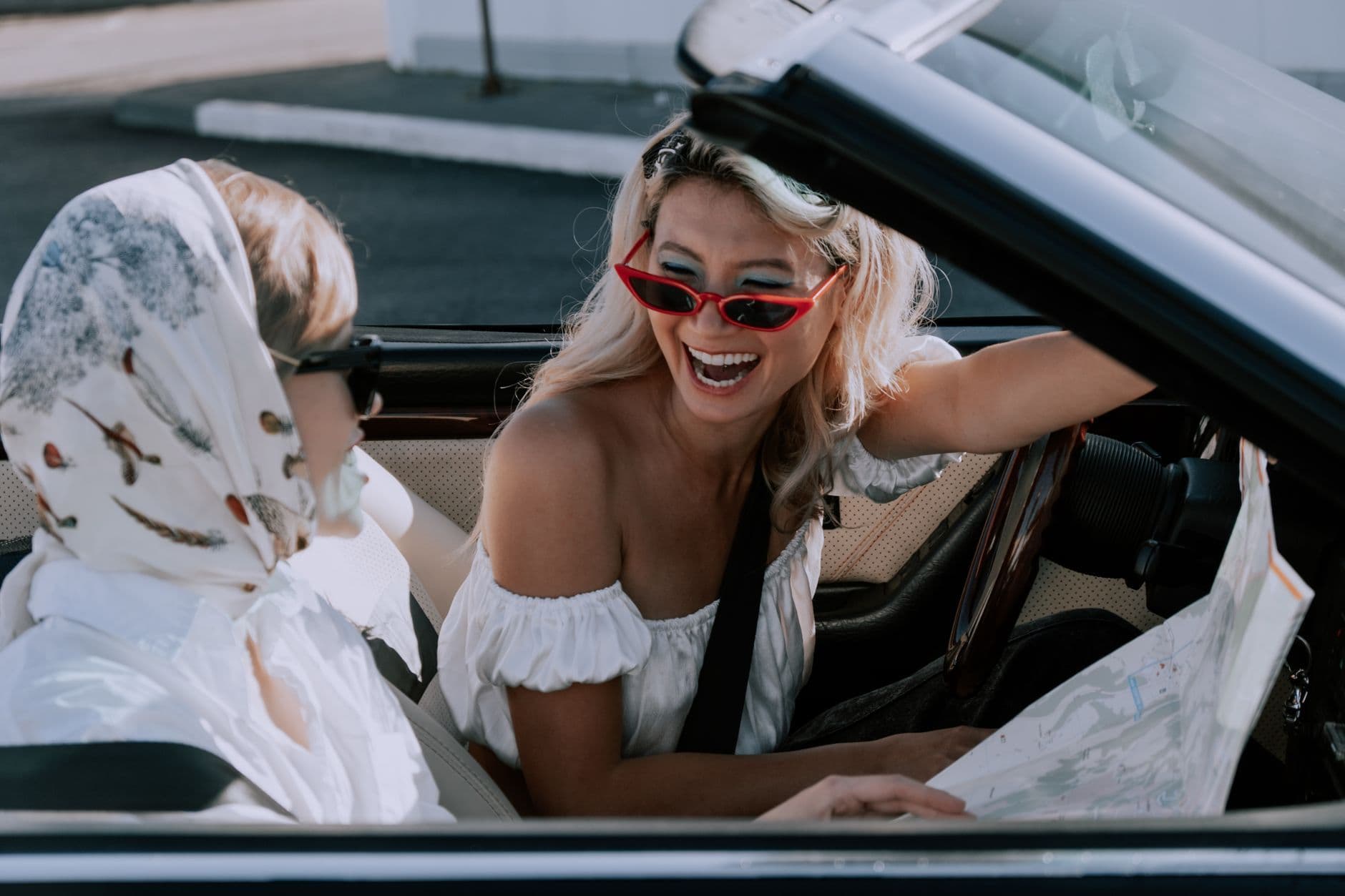 Two women laughing and enjoying a road trip in a convertible, holding a map.