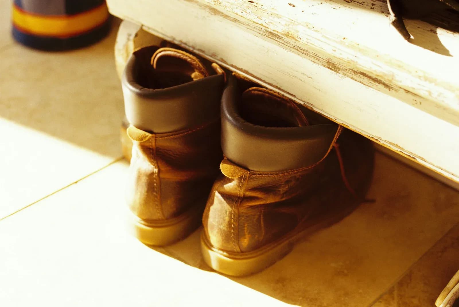 A pair of boots stored under a wooden bench in sunlight.